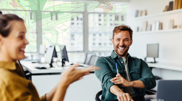 Zwei lachende Kollegen in einem modernen Büro mit Computern, im Hintergrund eine topografische Landkarte