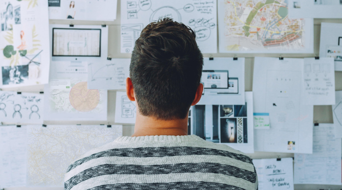 Person von hinten betrachtet eine Wand voller Pläne, Karten, Skizzen und Notizen – kreative Arbeitsumgebung oder Projektplanung