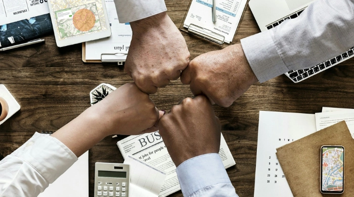 Vier Geschäftspartner stoßen mit Fäusten über einem Schreibtisch zusammen – Symbol für Teamwork, Vertrauen und erfolgreiche Partnerschaft bei onmaps, mit Karten und Geschäftsunterlagen im Hintergrund.